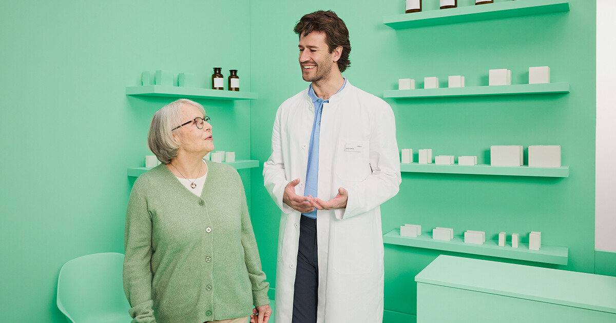 Apotheker und ältere Patientin unterhalten sich stehend in stilisierter Apotheke. 