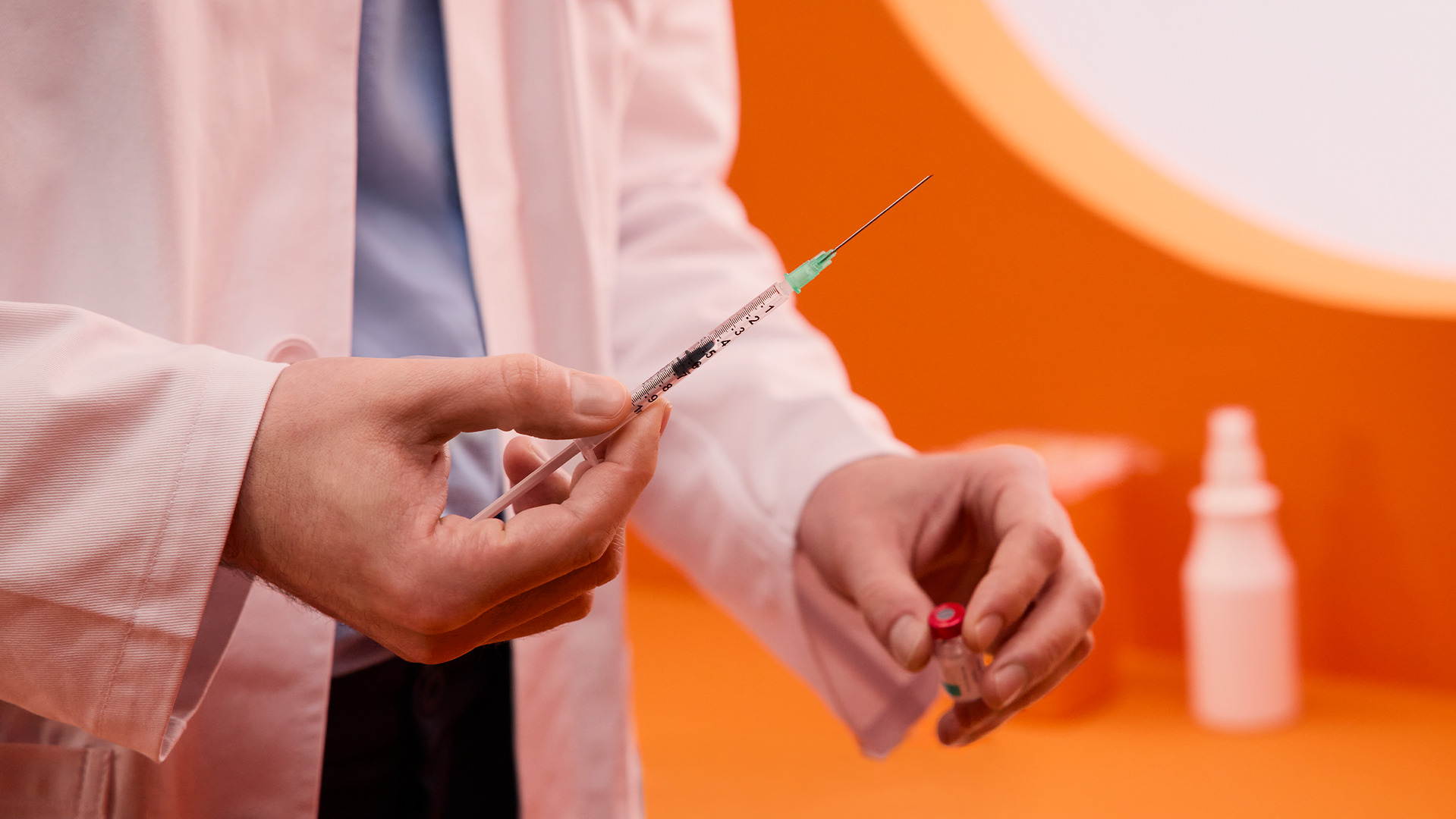 Hände eines Apothekers halten Spritze und Impfstoff-Vial in einem stilisiertem Impfzimmer hoch.